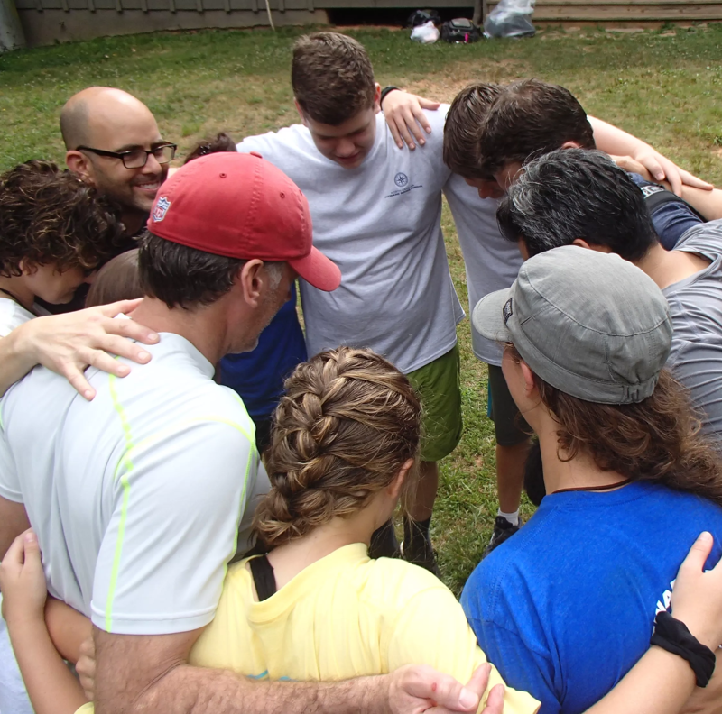 In an outdoor setting, a group of approximately ten people are gathered in a close circle, embracing each other in a group hug. The individuals vary in age and appearance, with some wearing casual clothing like t-shirts and hats. Their heads are bowed towards the center, suggesting a moment of unity, support, or shared emotion. The background features a grassy area and a glimpse of a building or fence.
